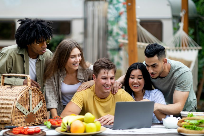 Group of multiracial millennial friends using laptop on autumn camping trip to countryside, chatting